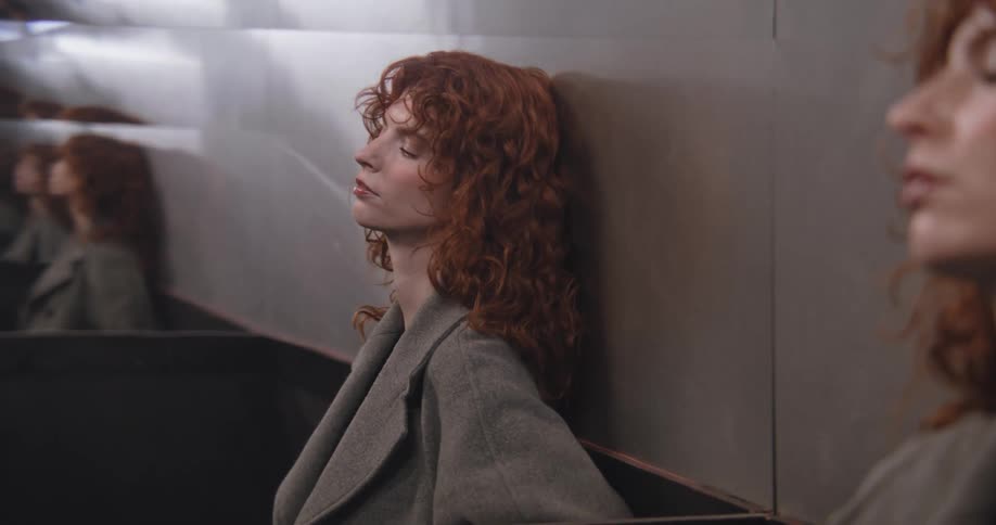 A melancholic red-haired woman in a grey coat leans against a mirrored wall, closing her eyes before looking into the camera with a sad expression