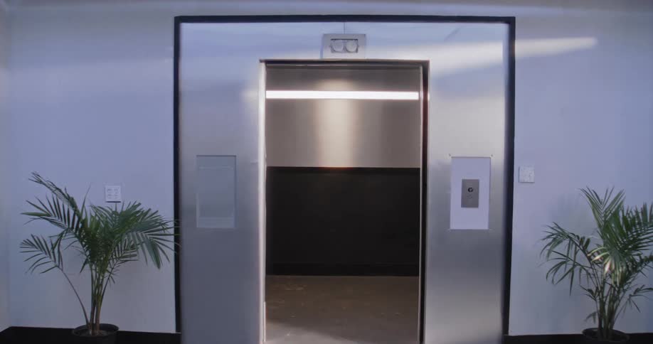 A slow camera push towards an open, empty elevator in a sterile and modern corporate lobby with potted plants