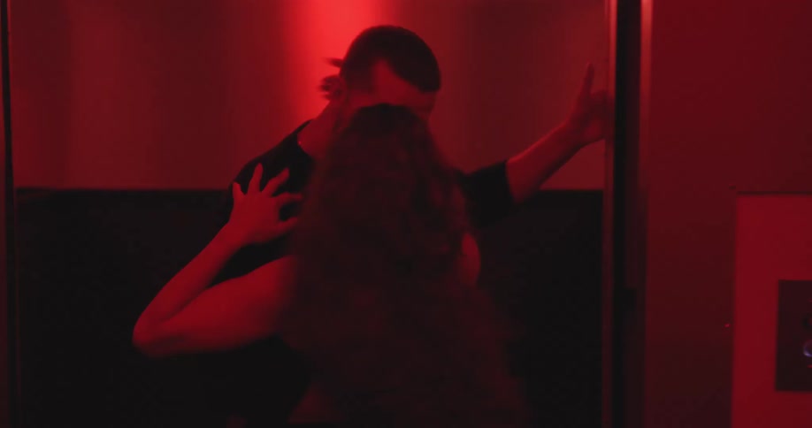 A passionate couple kisses as the elevator doors open, and the man carries the woman out into the red-lit hallway