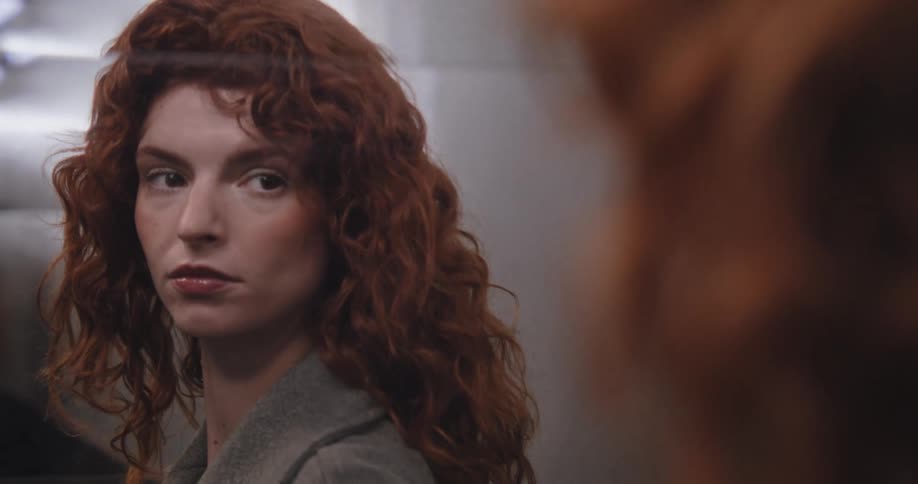 A serious woman with long curly red hair examines her reflection in a mirror with a look of unease