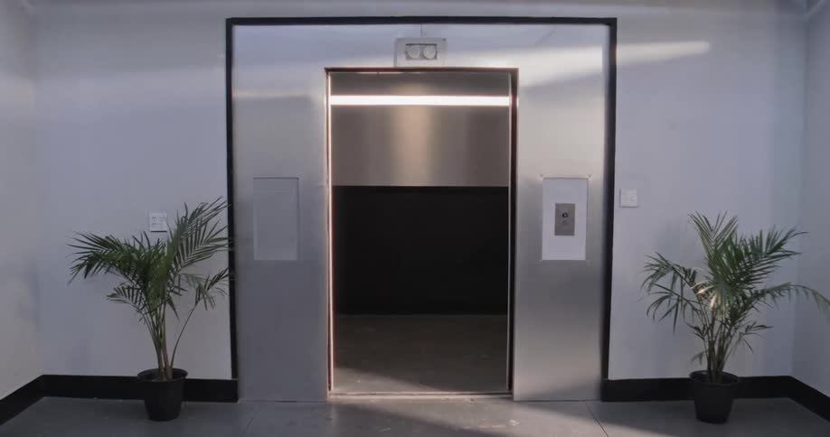 A slow dolly shot moves towards an open stainless steel elevator in a modern, sunlit building lobby