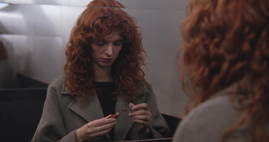 A beautiful woman with curly red hair gazes at her reflection while applying lipstick in a modern bathroom