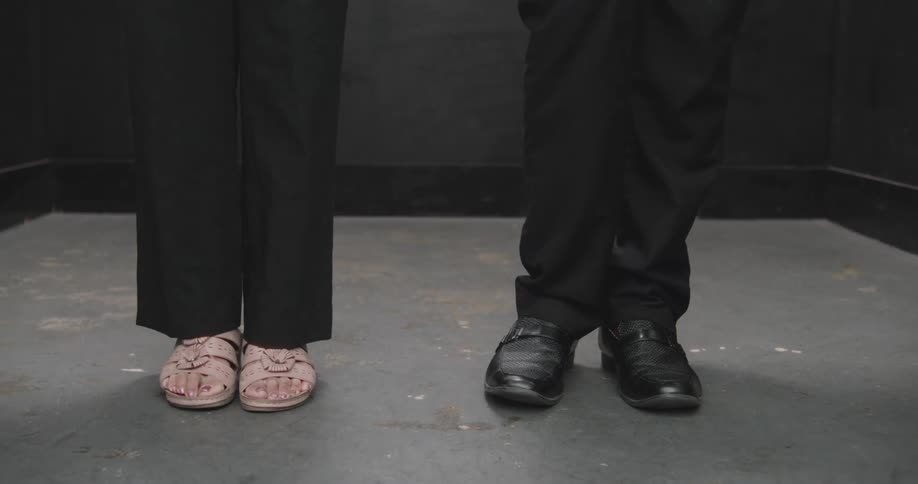 A close-up on the feet of a man and a woman wearing black pants, nervously tapping their shoes while waiting
