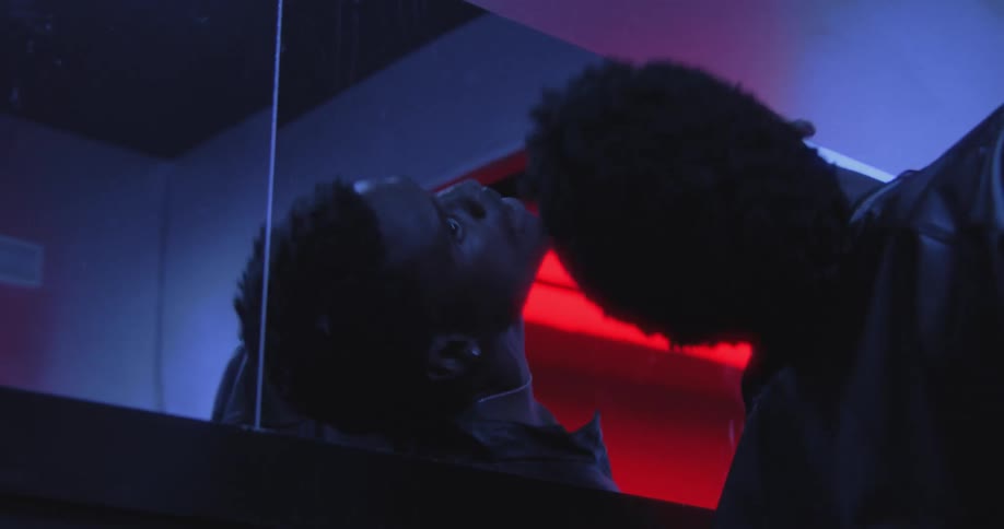 A young man has a dramatic psychological breakdown while looking at his reflection in a mirror under intense blue and red lighting