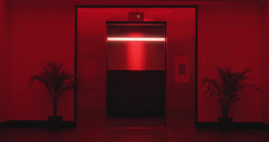 An elevator door opens and then closes in a dark, ominous hallway bathed in intense red light