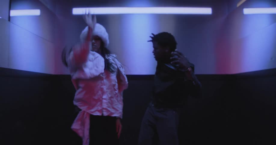 An elevator door opens in a red hallway to reveal a stylish couple energetically dancing inside