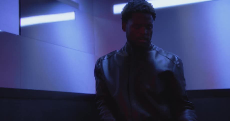 A stylish young man dances expressively in a modern elevator illuminated by cool blue and vibrant red lighting
