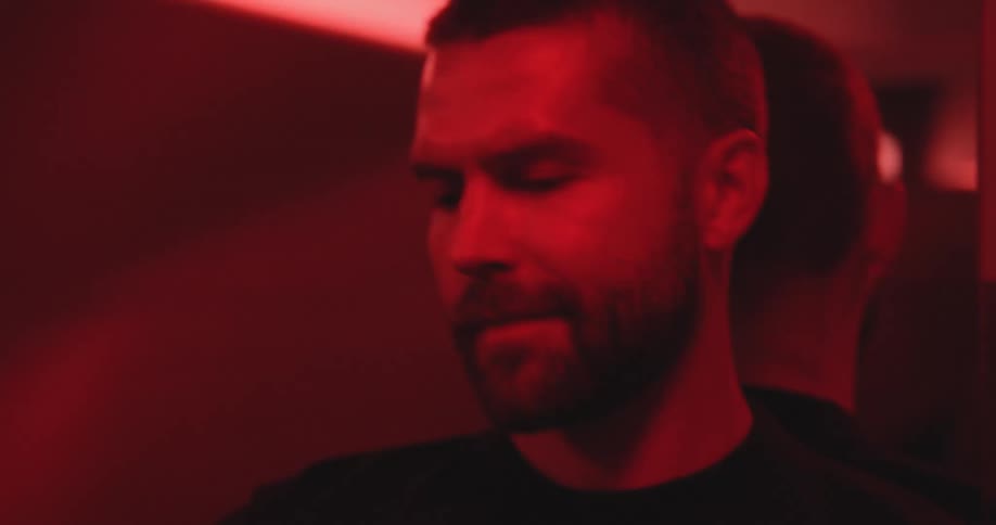 A handsome bearded man smiles at a woman as they stand together in a dimly lit room with moody red lighting