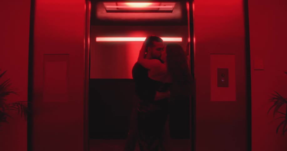 A passionate couple kisses and embraces as they emerge from an elevator bathed in intense red light