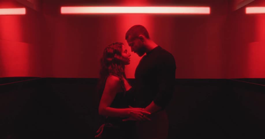 A passionate couple shares a romantic kiss in a red-lit elevator as the doors close on their intimate moment