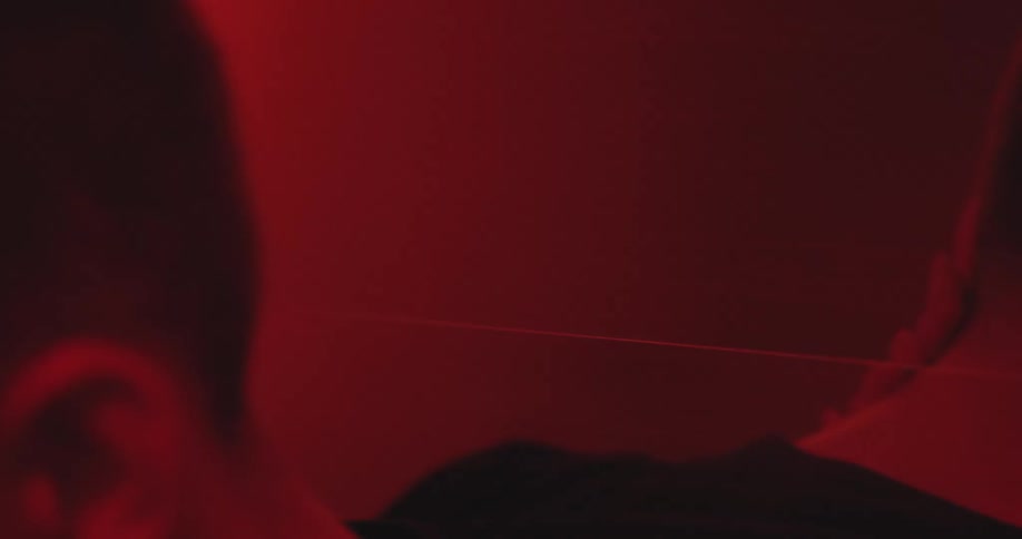 A woman runs her hands through a man's hair during a passionate embrace under dramatic red lighting