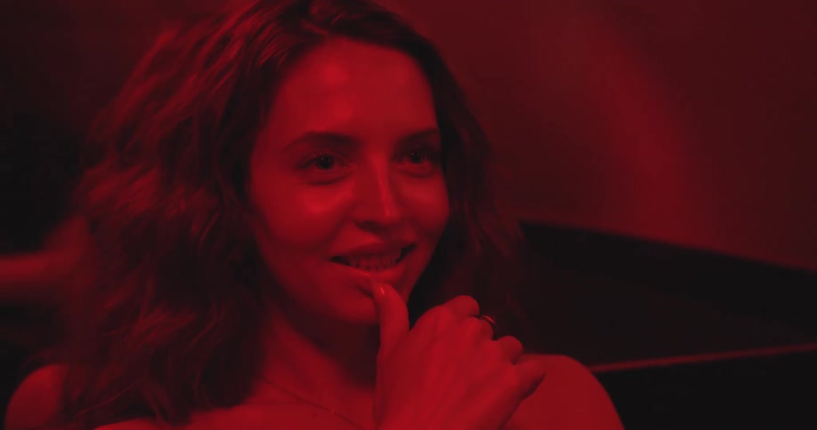 A sensual young woman looks on with a playful smile, her face illuminated by a dramatic red light