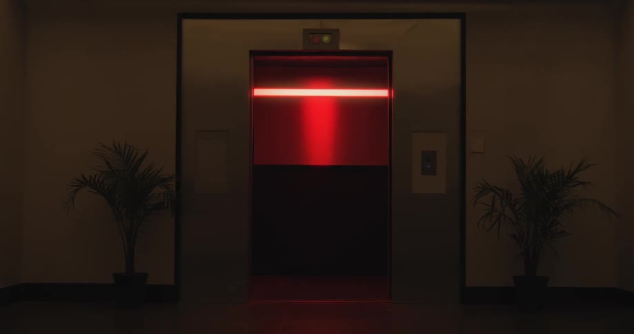 A sinister red light glows from within a dark elevator as its doors open and close in a creepy hallway