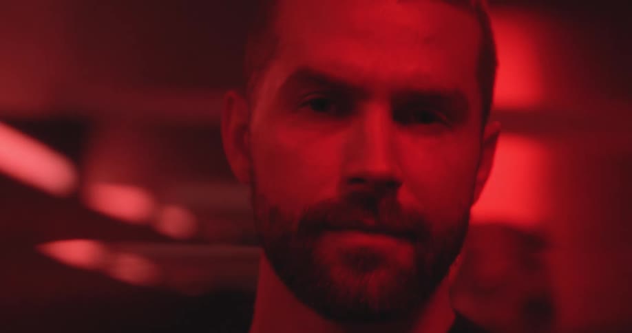 A bearded man gives an intense and serious stare directly into the camera in a dark room bathed in red light