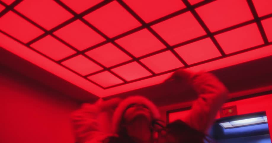 A stylish man and woman perform an expressive and dynamic dance routine in a surreal hallway illuminated by intense red lighting