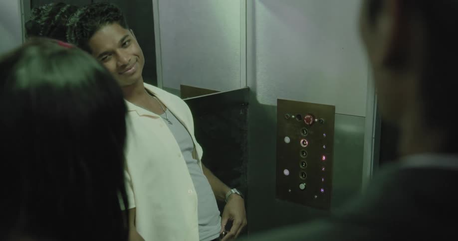 A young man playfully presses all the buttons on an old elevator panel while riding with friends