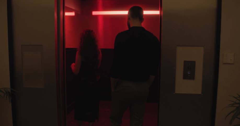 A stylish woman in a black dress calls an elevator and is joined by a man before the doors open to reveal a dramatic red-lit interior