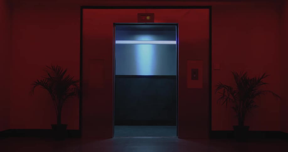 In a dark, red-lit corridor, modern elevator doors slowly open and then close again