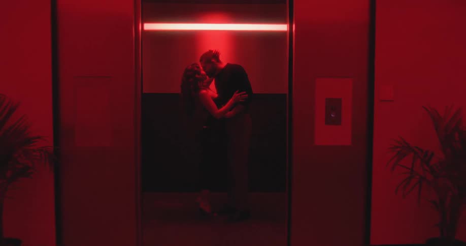 A silhouetted couple passionately kisses in a red-lit elevator as the camera pulls back and the doors slowly close