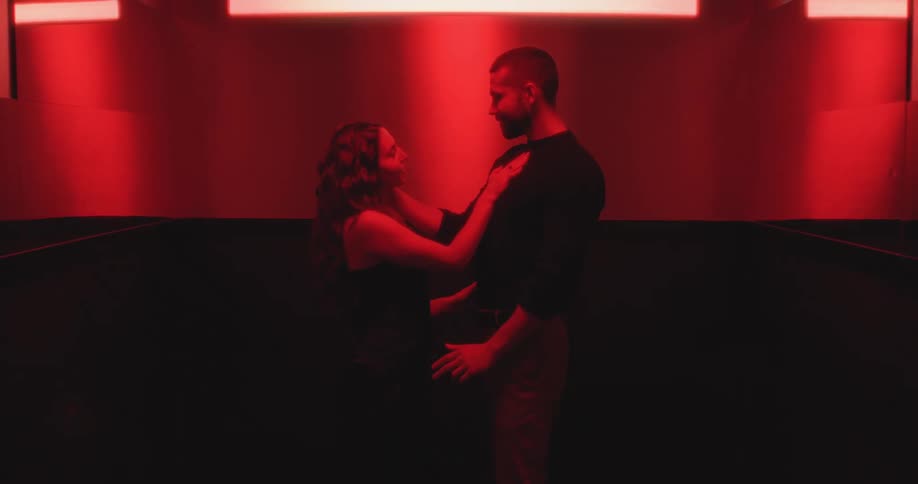A passionate couple slowly embraces and shares an intimate kiss in a dark, red-lit room