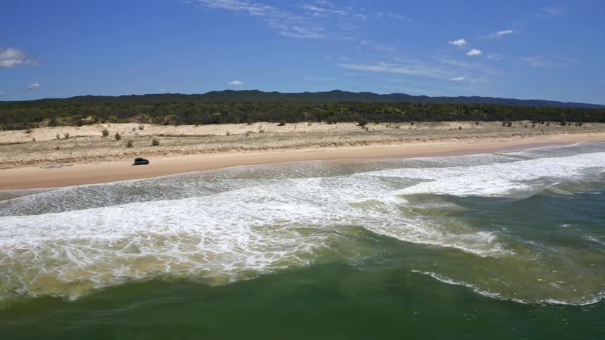 An epic aerial view captures an SUV cruising along a remote, sandy beach as gentle ocean waves roll ashore