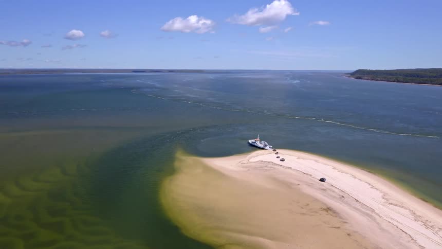 An aerial view shows a vehicle ferry docked at a long sand spit, with cars waiting to cross a wide channel on a sunny day
