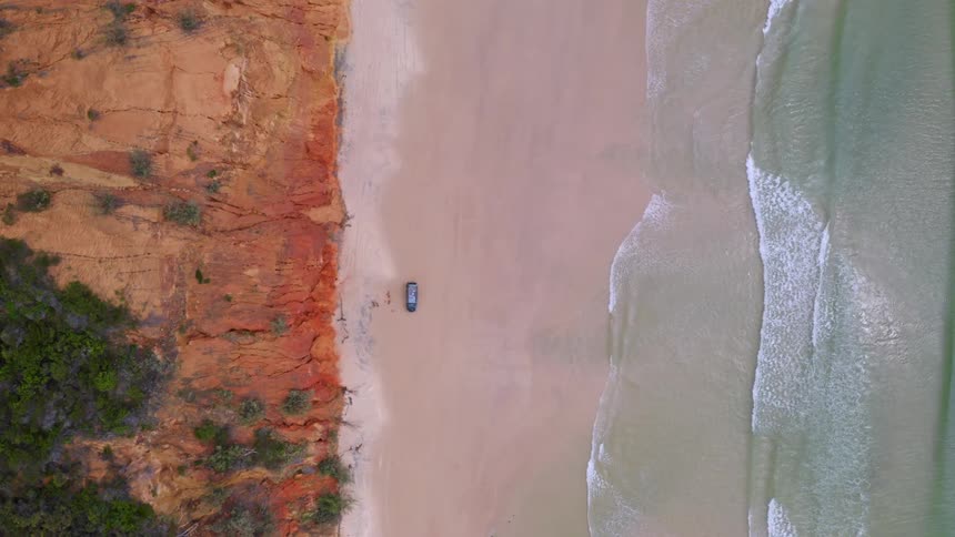 A lone SUV is parked on a secluded beach, captured in a static top-down aerial shot showing the contrast between the red earth cliffs and the gentle ocean waves