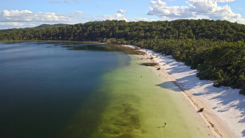 An expansive aerial shot slowly pulls back to reveal a picturesque freshwater lake with brilliant white sand shores surrounded by a lush green forest