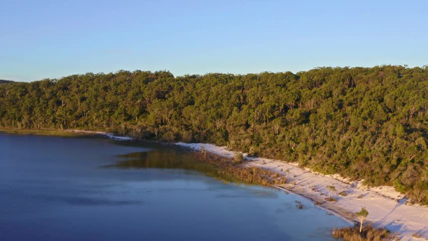 Aerial view of a serene blue lake surrounded by a dense forest and a white sandy shore during the golden hour
