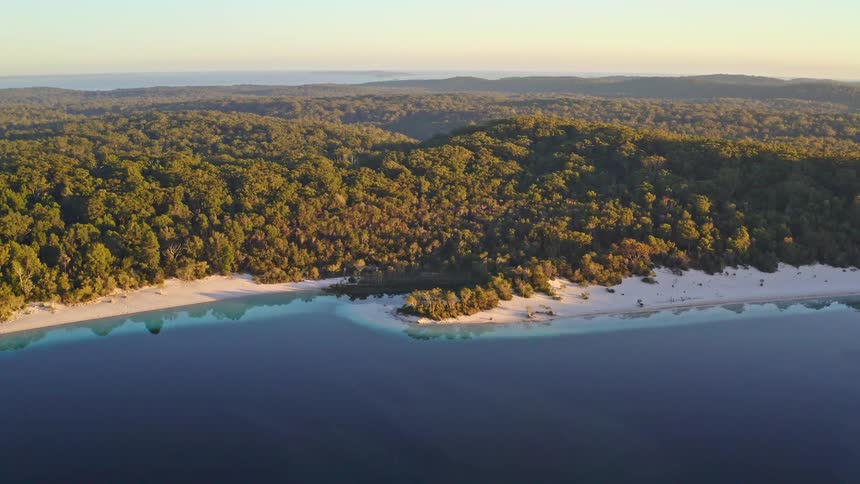 A beautiful static aerial shot captures the golden hour light over a vast green forest and a white sand beach bordering a pristine blue lake