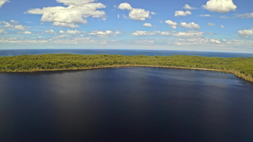 A stunningly still aerial cinemagraph captures a serene blue lake surrounded by a dense forest, with the distant ocean visible under a partly cloudy sky