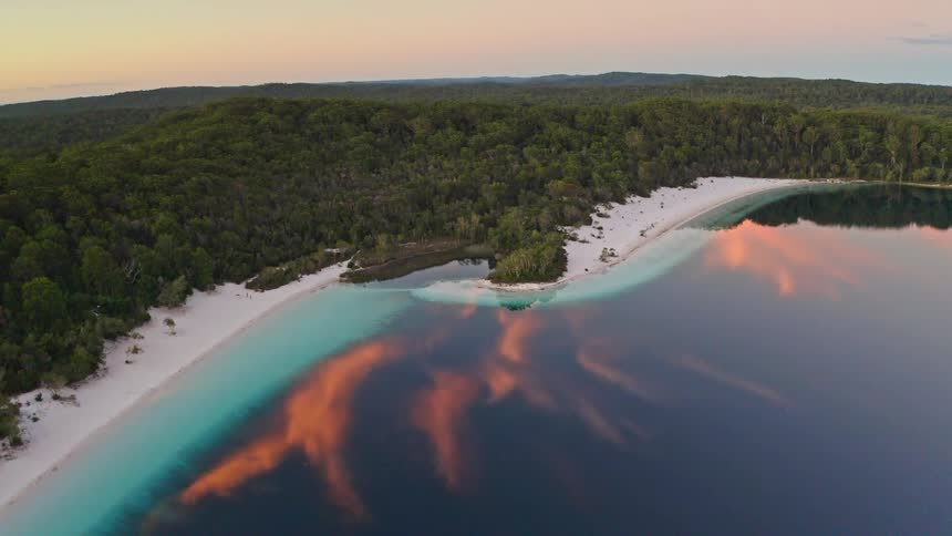 A tranquil aerial view shows a white sand beach bordering a calm lake and dense forest, with a colorful sunset reflected in the water