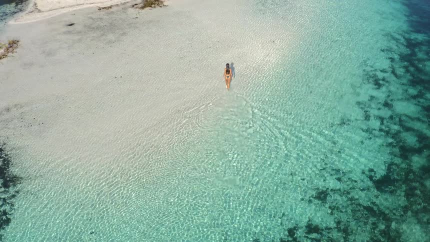 An aerial view reveals a woman in a bikini walking through the crystal-clear turquoise water of a beautiful secluded lake towards a white sand island