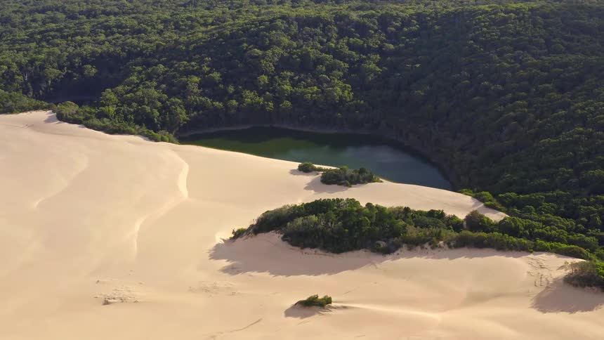 A spectacular aerial shot pulling back to reveal a vast sand dune system encroaching on a dense green forest and a tranquil freshwater lake