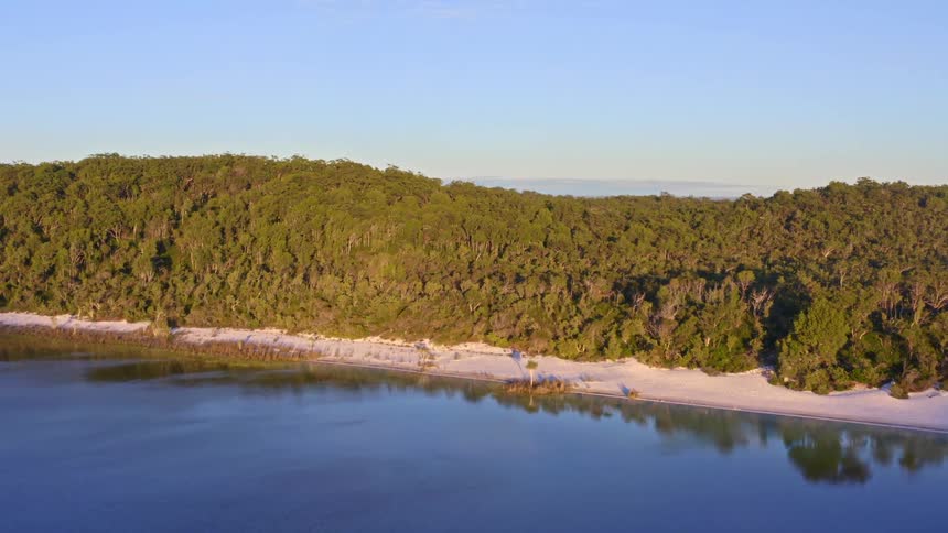 A slow aerial shot tracks along a pristine white sand beach bordering a tranquil blue lake and a dense green forest at sunrise