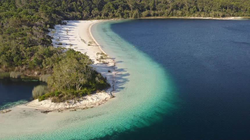 Stunning aerial reveal of a pristine lake with a white sand beach, separating shallow turquoise water from the deep blue