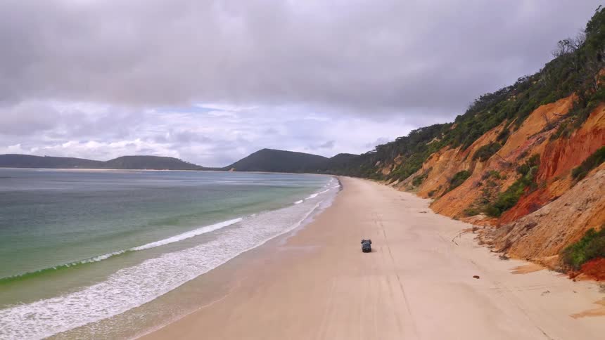 An aerial view shows a 4x4 vehicle driving along a vast, empty beach next to colorful cliffs and the ocean on an overcast day