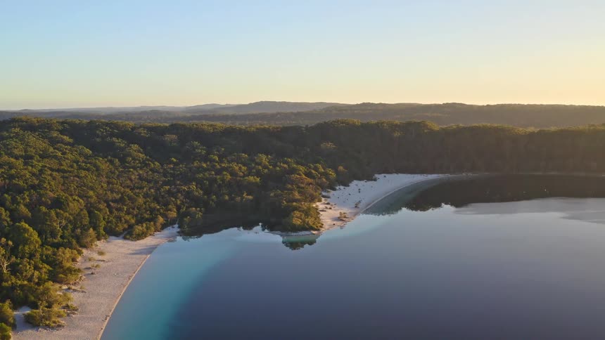 A stunning aerial view of a pristine blue lake with a white sand beach nestled in a vast, lush forest at golden hour