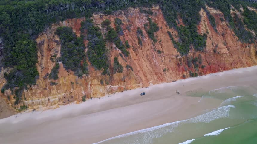 A slow aerial reveal shot of a solitary 4x4 vehicle on a vast beach, framed by the ocean and stunning, colorful sand cliffs topped with a dense forest