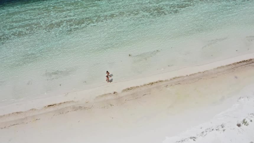 Slow motion aerial shot zooms in on two women in bikinis as they walk along the shore of a beautiful white sand beach with turquoise water