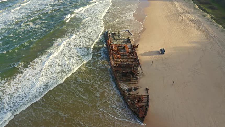 A high-angle aerial view captures the immense Maheno shipwreck resting on a sandy beach, with gentle ocean waves lapping at its rusted hull