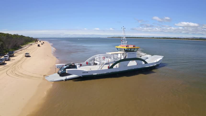 An aerial view captures a 4x4 vehicle driving off a car ferry that has landed on a beautiful sandy beach