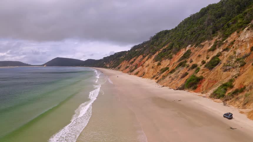 A scenic aerial drone shot captures 4x4 vehicles driving along a vast, sandy beach flanked by colorful, forested cliffs on an overcast day