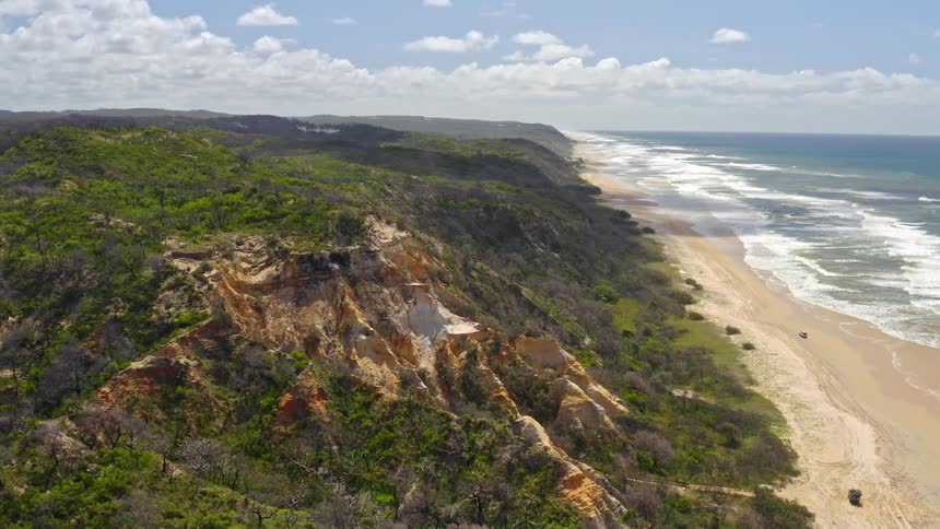 A slow-moving aerial shot reveals the dramatic and colorful sand cliffs of a remote coastline next to the ocean on a beautiful sunny day