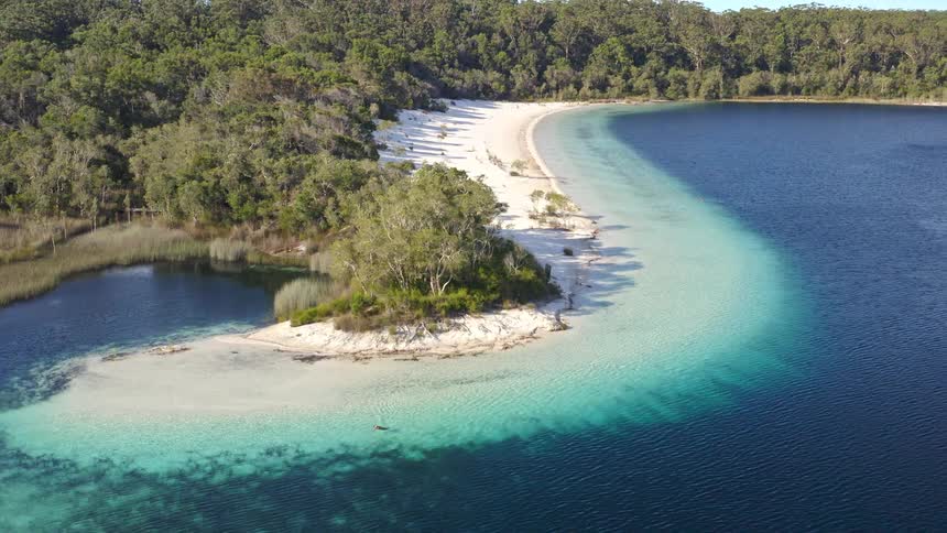 An idyllic aerial shot captures a pristine freshwater lake with a white sand beach curving through crystal clear turquoise waters, surrounded by a dense forest