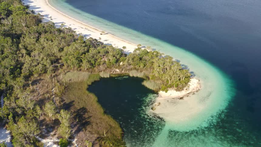 An incredible aerial pullback shot revealing a pristine island landscape featuring a freshwater lake with turquoise shallows, deep blue water, and a white sand beach