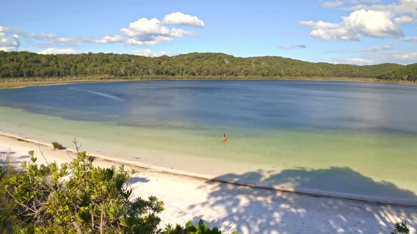 A slow drone dolly shot revealing a lone person wading in the pristine, clear waters of a lake with a white sand beach