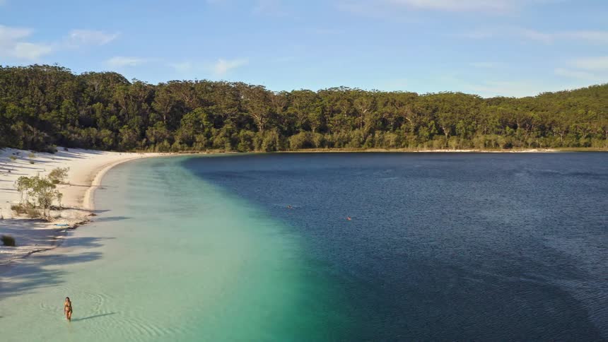 An aerial view pans across a stunning lake with a white sand beach, revealing a sharp contrast between shallow turquoise and deep blue water