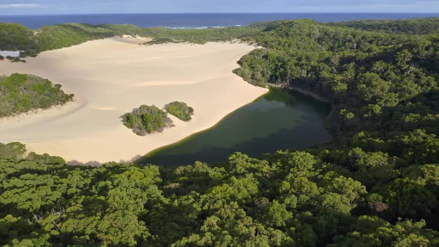 A slow aerial pullback reveals the stunning Lake Wabby on Fraser Island, a unique freshwater lake bordered by a massive sand dune and a lush forest