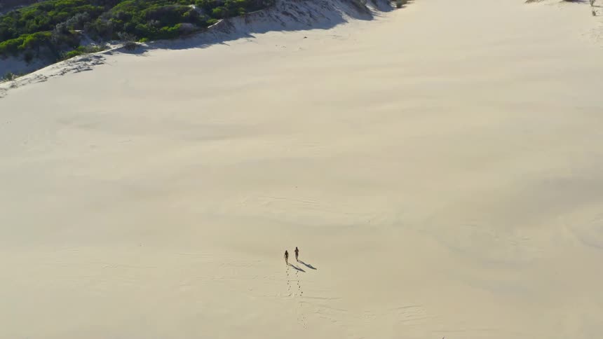 An aerial view pulls back to reveal a man and woman walking across expansive sand dunes towards the coast